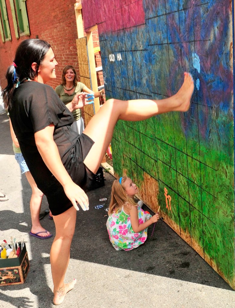 FANCY FOOTWORK: Madison Boynton, bottom, watches as Deanna Thibeau pulls her leg back after making a blue-painted footprint on a plywood mural wall Wednesday in Dummers Lane. The mural on the temporary plywood wall in the alley between the Liberal Cup and Higher Grounds will be part of the Old Hallowell Day beer garden at Higher Grounds.