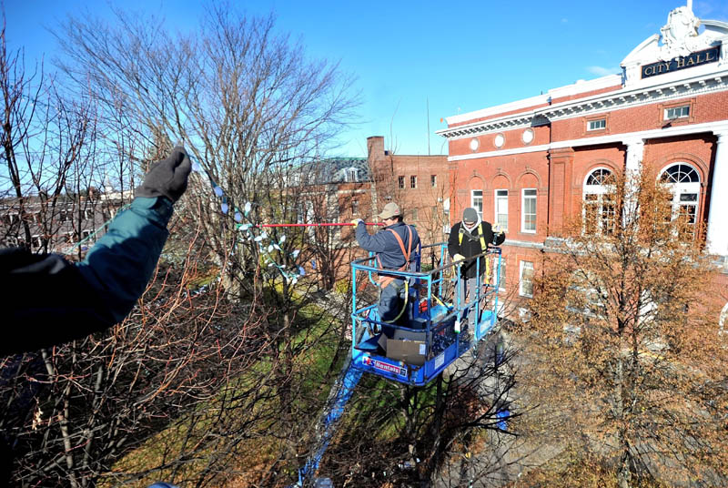 Staff photo by Michael G. Seamans City Hall employee, Dave Higgins, far left, holds a strand of lights as public works employee, Mike Folsom, center, guides them on to the tree from a cherry picker with City Hall employee Lance Bates, right, who is untangling a strand as they decorate the trees in Castonguay Square in Waterville for the holiday season on Friday morning.