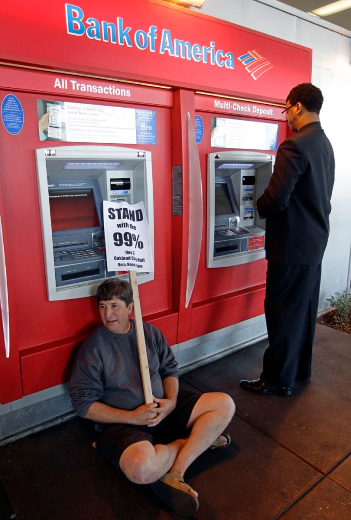 DEMANDING CHANGE: A protester sits in front of an ATM as a customer gets money Wednesday at a Bank of America branch in Oakland, Calif. The spirit behind Bank Transfer Day caught fire with the Occupy Wall Street protests around the country and had more than 77,000 supporters on its Facebook page as of Friday. The movement helped beat back Bank of America’s plan to start charging a $5 debit card fee.