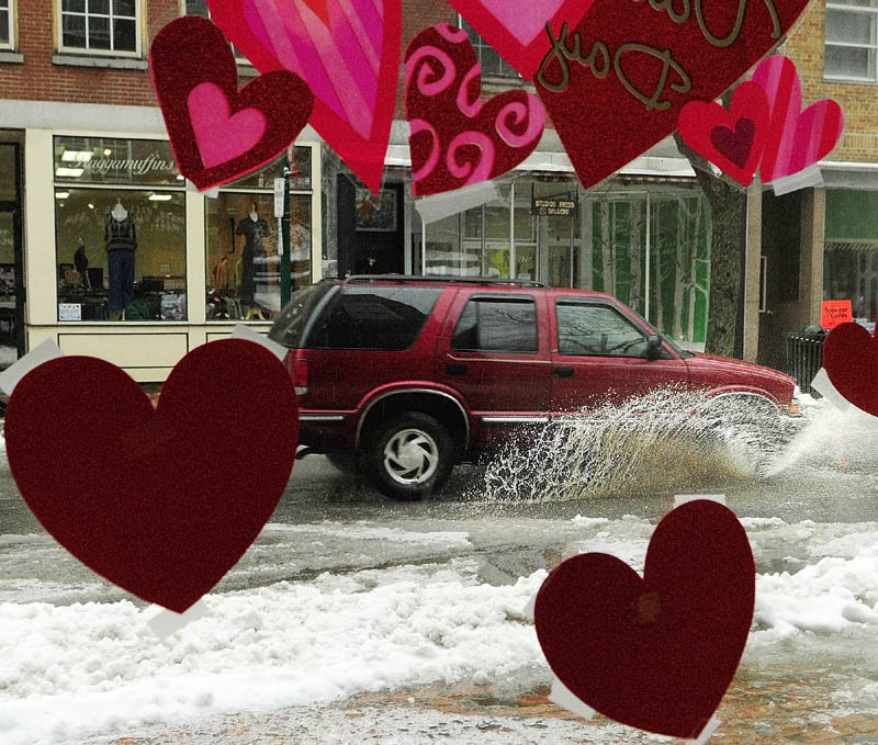 Valentine’s Day decorations in the Johnson Hall Performing Arts Center office window frame a vehicle splashing down Water Street in downtown Gardiner on Friday afternoon. The storm had started as snow and switched to sleet and rain as the day went on.