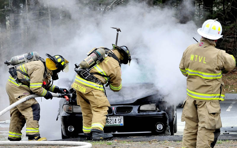A Waterville firefighter sprays water on a burning vehicle while another firefighter uses an axe to raise the hood of the car parked at Thomas College in Waterville on Wednesday.