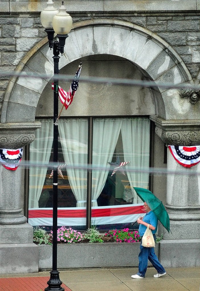An umbrella-wielding pedestrian walks past bunting-draped window and pillars on Friday in front of the Olde Federal Building in Augusta. The large, granite office building is at the corner of Winthrop and Water Streets in downtown Augusta.
