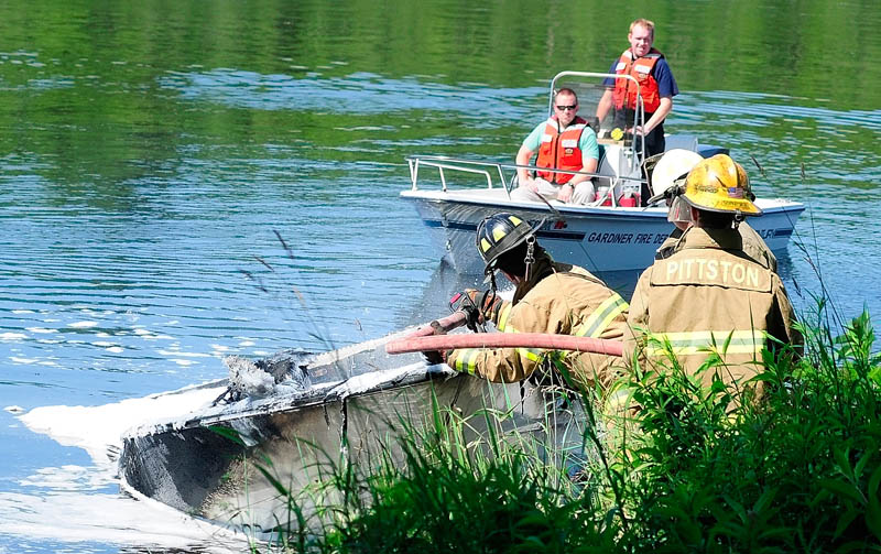 A Gardiner fireboat floats past as firefighters on shore spray foam on a burned boat on Friday, on the banks of the Kennebec River near Togus Stream in Pittston. Four people were on the boat when a fuel vapor explosion caused the fire, according to fire officials. The boat drifted down river from Gardiner landing to point where firefighters extinguished flames.