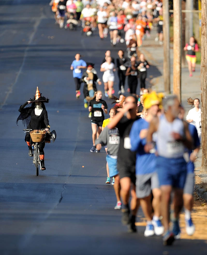 Laura Patterson, left, rides her bike dressed as a witch as nearly 200 runners file down Mayflower Hill Road during the Freaky 5K Fun Run that starts and stops at Colby College organized by Hardy Girls Healthy Women today.