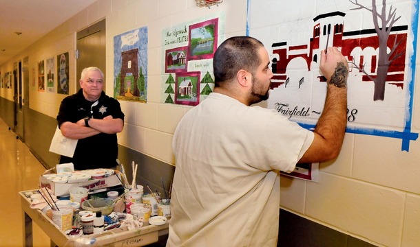 LOCAL POSTCARDS: Somerset County Jail inmate Leo Coutu adds color to one of 28 images from the Somerset Bicentennial quilt of area town landscapes on the walls at the Madison facility on Wednesday. Watching is Compliance Officer Sean Maguire. Plans are underway to transfer the scenes into postcards as part of a prison industry.