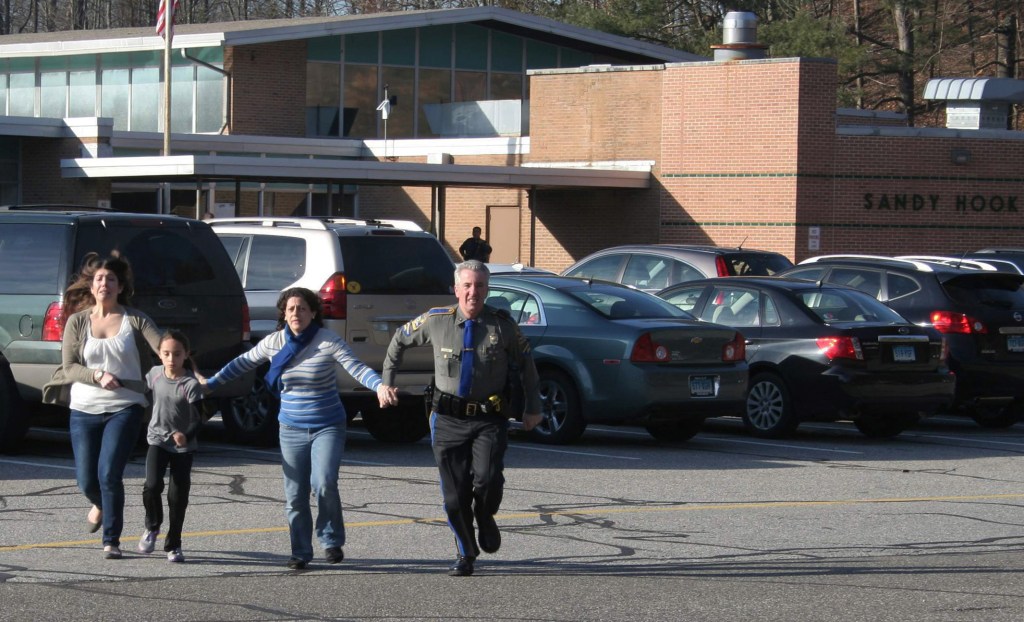 FILE - In this Friday, Dec. 14, 2012, file photo provided by the Newtown Bee, a police officer leads two women and a child from Sandy Hook Elementary School in Newtown, Conn., where a gunman opened fire, killing 26 people, including 20 children. A prosecutor is planning to release a report on the investigation into the massacre at Sandy Hook Elementary School on Monday, Nov. 25, 2013, but the public will have to wait longer for the Connecticut state policeís full accounting of the crime.(AP Photo/Newtown Bee, Shannon Hicks, File) MANDATORY CREDIT: NEWTOWN BEE, SHANNON HICKS