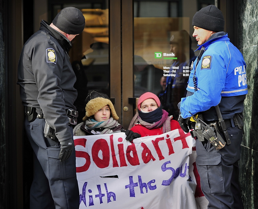 Elizabeth Catlin of Brunswick, seated left, and Seth Schlotterbeck, also known as Sylvia Stormwalker, of Auburn attached themselves with bicycle locks around their necks to the door handles of TD Bank on Congress Street in Portland in a protest against investment in the Keystone XL pipeline. Portland police officers Dave Argitis, left, and Dan Knight stand by them as other officers try to remove the handles from inside the bank on Wednesday.