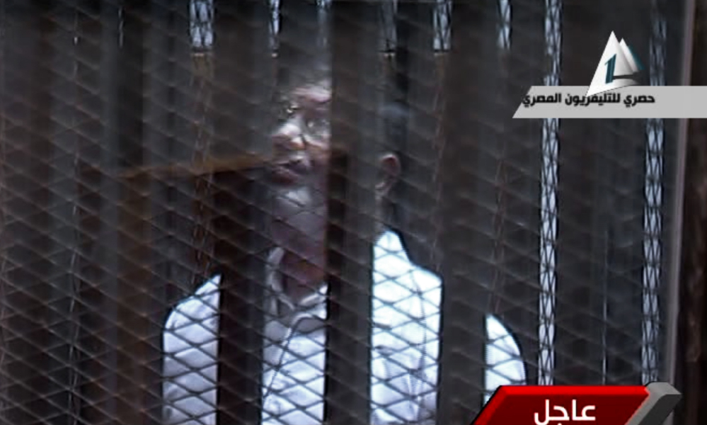 In this image taken from Egypt State TV, Egypt’s toppled President Mohammed Morsi stands inside a glass-encased metal cage in a courtroom in Cairo on Tuesday.