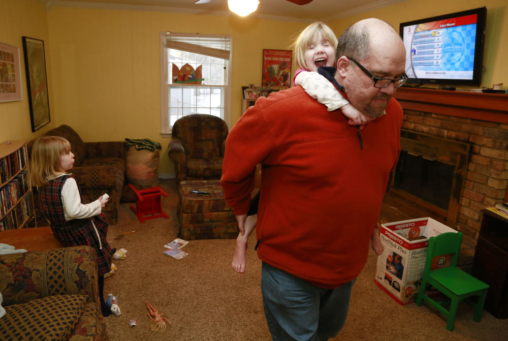 Mike Beck carries his daughter Veronica on his back through the living room as his daughter Maria, left, plays a video Monday.