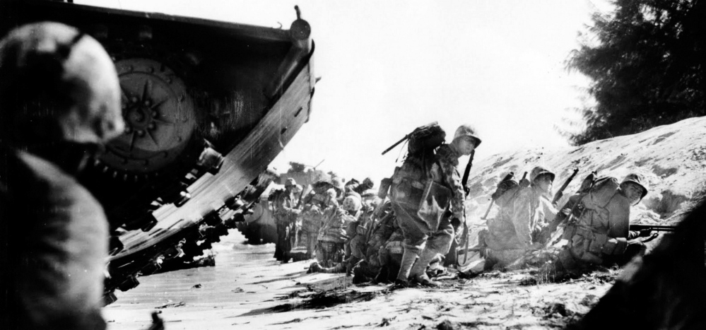 ON THE BEACH: The first wave of Marines takes cover behind the sand dunes on Saipan beach, during the World War II invasion of Marianas Islands. The soldier kneeling in the sand at far right is the essay’s author, Carl Matthews of Texas; second from right is Wendal Nightingale of Skowhegan, Maine; standing is Lt. James Leary of North Carolina. Neither Nightingale nor Leary made it home from Saipan; both are still listed as missing in action.