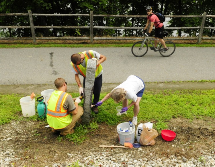 A passing cyclist watches city workers Taylor Perry, left, and Shawn Winslow and volunteer Kathleen “Sukey” Sikora remove graffiti from a Kennebec River Rail Trail milestone on Wednesday in Augusta.