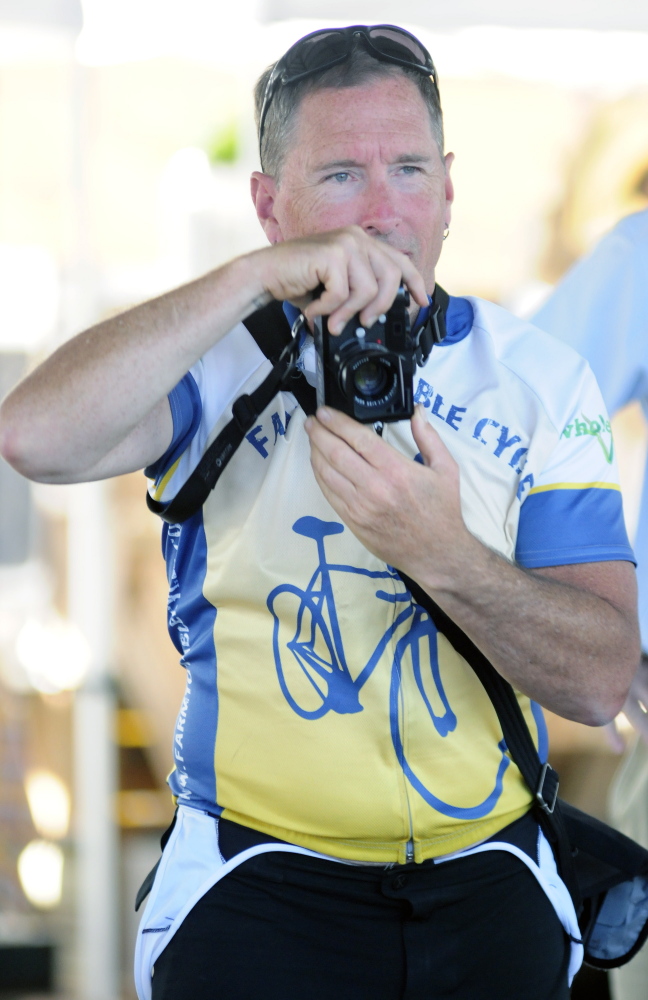 Glenn Charles looks over the top of his camera while photographing speakers Tuesday during an event at the farmers market at Mill Park in Augusta. He is on a bicycle tour to learn about food in New England.