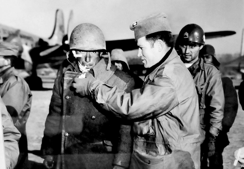 A photograph by Galen Leavitt shows an officer lighting the cigarette of a wounded soldier during the Korean War.