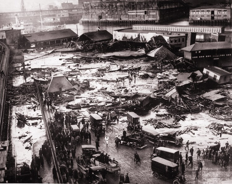 Twenty-one people were killed when a tank of molasses ruptured and exploded, sending a 40-foot wave of the liquid down Boston's Commercial Street at a speed of 35 mph. Wreckage of the collapsed tank is visible in the background, center, next to the light-colored warehouse. The elevated railway structure is visible at far left and the North End Park bathing beach is to the far right. Boston Public Library photo
