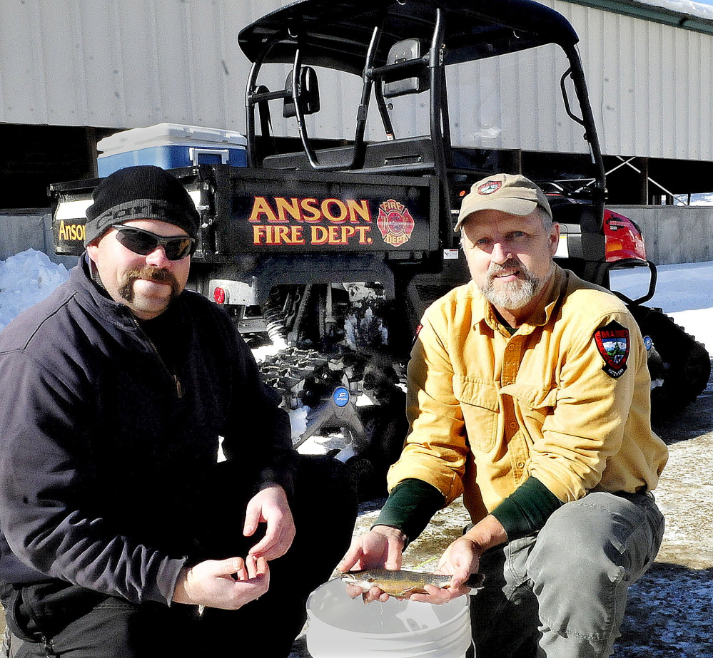 Anson Fire Chief Jeremy Manzer, left, and Gene Arsenault, of the Embden Rearing Station, hold a tagged brook trout that is worth $5,000 if caught by a participant in this Saturday’s fishing derby on Embden Pond. Behind the men is a Fire Department all-terrain-vehicle bought last year.