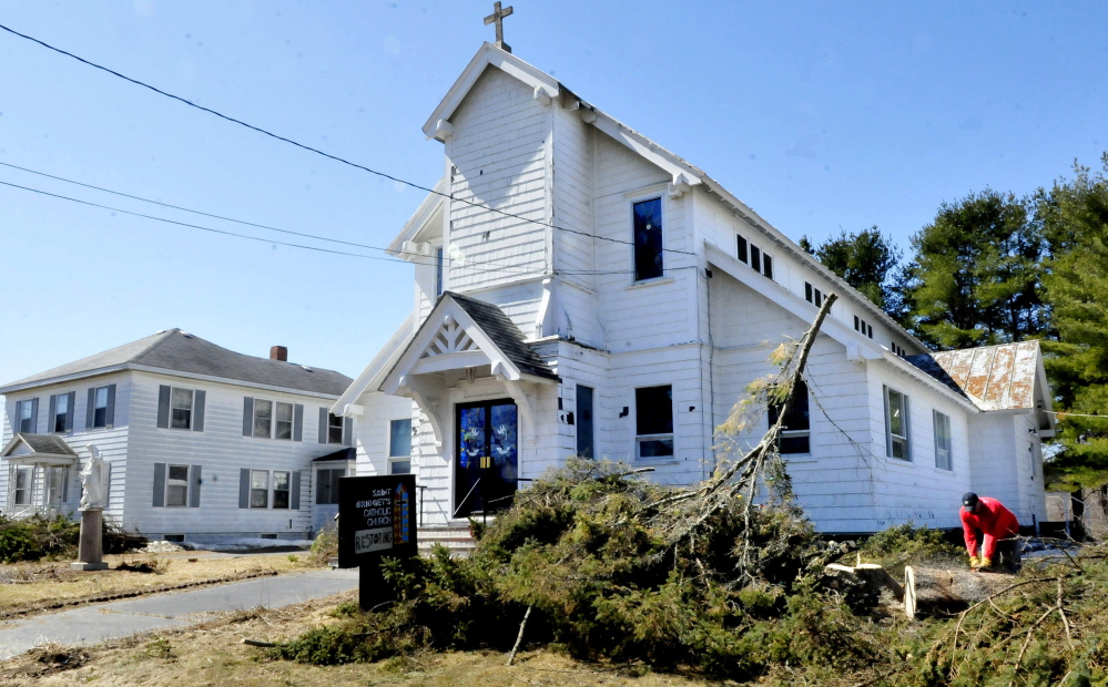Robert Searles cuts up a tree he cut down beside St. Bridget’s church in Vassalboro on Sunday. Rachel and James Kilbride bought the closed church and rectory, left, and plan to restore the buildings and open them as an event hall and community center.