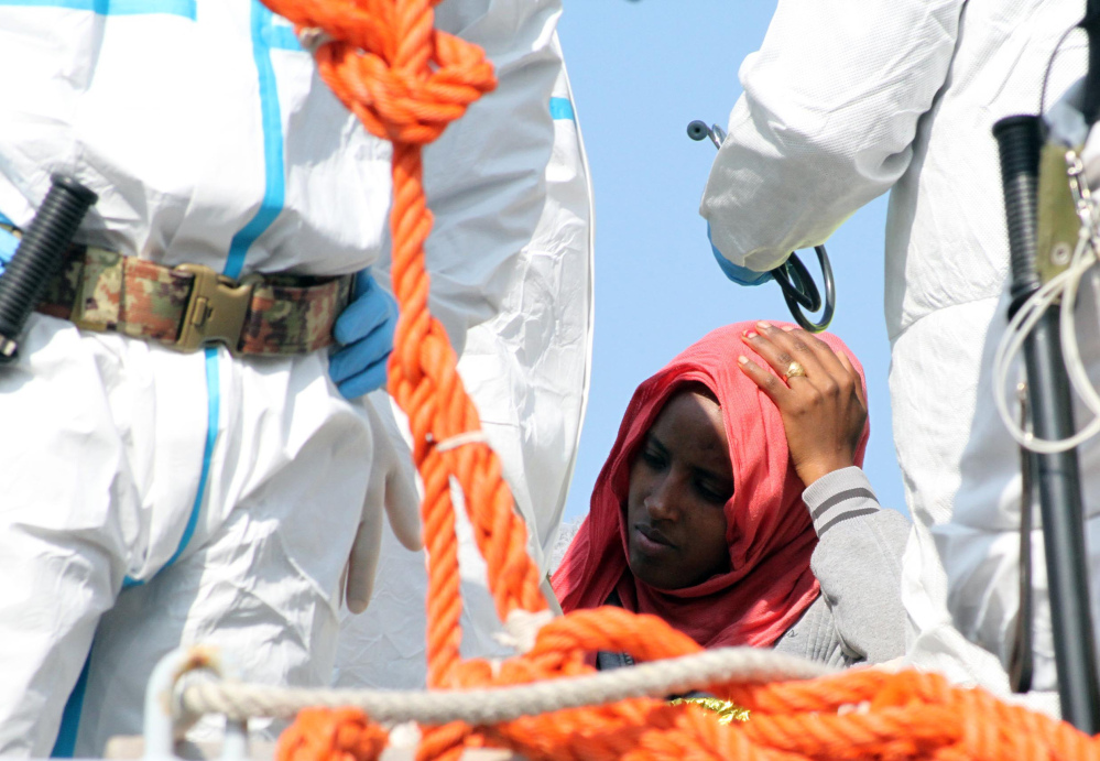 A woman waits to disembark from an Italian Navy vessel in southern Italy after being rescued in the Mediterranean, as workers in protective jumpsuits process incoming migrants.