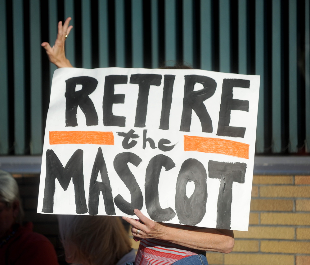 A woman holding a sign urging Skowhegan to change the mascot at the local high school waves on Thursday to a supporter in Skowhegan.