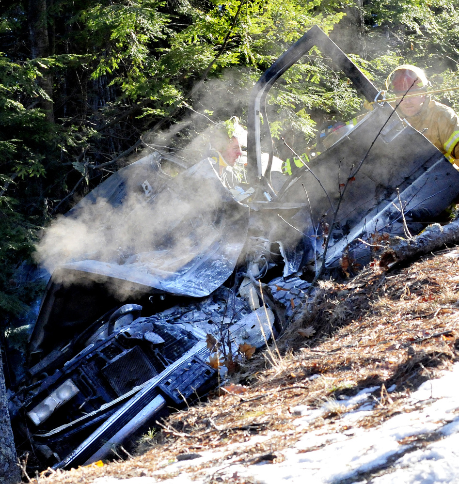 As firefighters stabilize an injured man inside the vehicle, smoke continues to rise 30 minutes after it went off Route 135 in Belgrade and slid down a steep embankment on Tuesday.