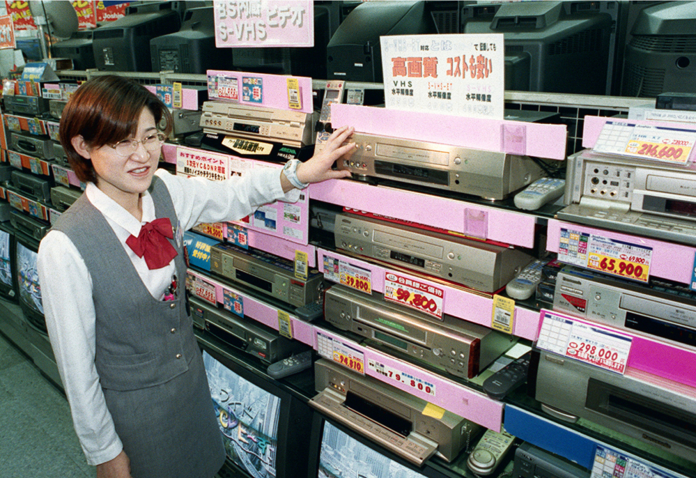 In this 1998 photo, a salesclerk shows high quality VHS video casette recorders at a home and electrical appliance store in Osaka. Japanese electronics maker Funai Electric Co. says it's yanking the plug on the world's last video cassette recorder. A company spokesman, who requested anonymity citing company practice, confirmed Monday that production will end sometime this month, although he would not give a date.