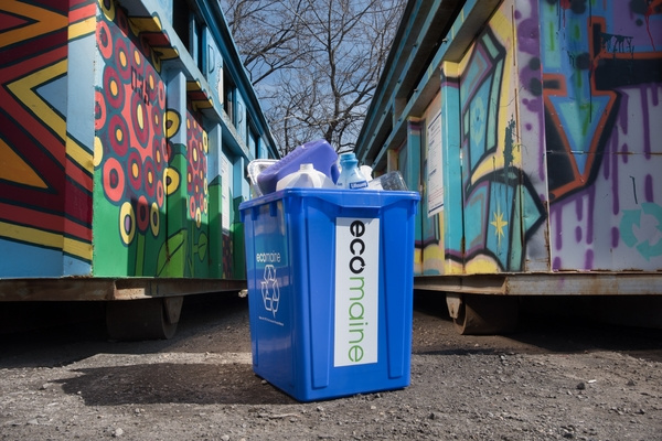 A single-sort recycling bin is seen in front of a painted recycling drop-off location for ecomaine.