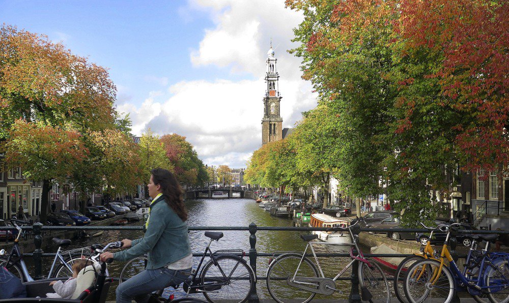 A woman rides her bicycle across a bridge over the tree-lined Prinsengracht canal in the center of Amsterdam. Trees play a critical role in urban environments, helping keep cities cool, mitigating air and noise pollution, and just making them more pleasant places to live and work.