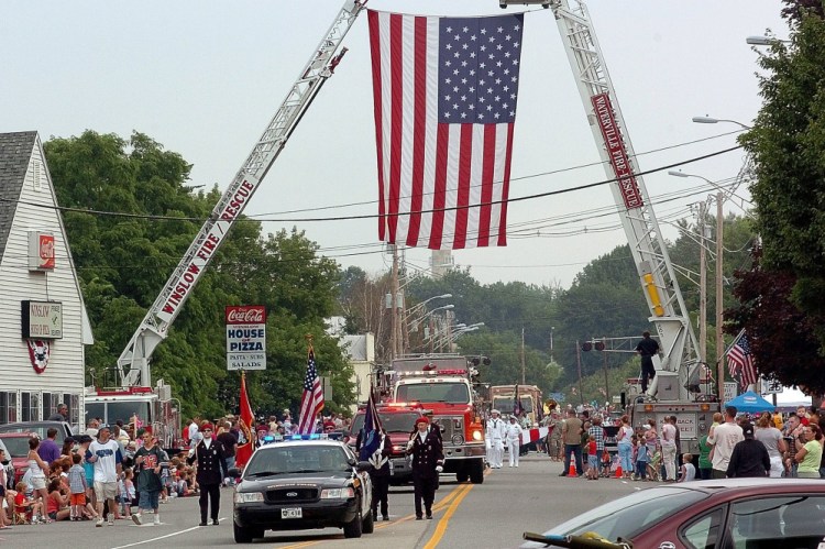 Participants of a previous Winslow Fourth of July parade pass under a huge American flag on Bay Street suspended by ladder trucks from the Winslow and Waterville fire departments. Event organizers aren't sure where the celebration will be held this year, if at all.
