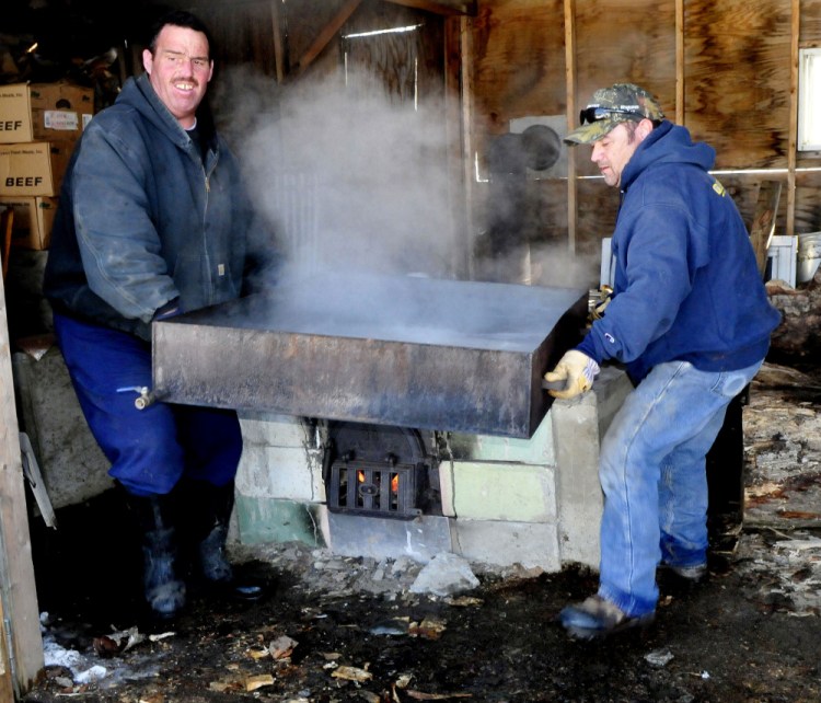 Paul Elkins, left, and Kurt Parker lift a tray of boiling maple syrup off a fire pit to pour into containers at their sap house in Thorndike on Thursday. Elkins said that, weather permitting, the pair and neighbor Ray Peabody will tap 60 trees and produce 25 gallons of syrup. "This is a family tradition that I've always done," Elkins said.