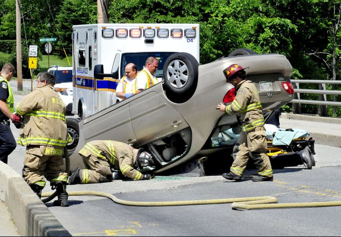 Waterville firefighters, including Chief Dave Lafountain lying on pavement at right, remove the driver of this vehicle that overturned in the middle of Thayer Memorial Bridge on Gilman Street in Waterville on Monday.