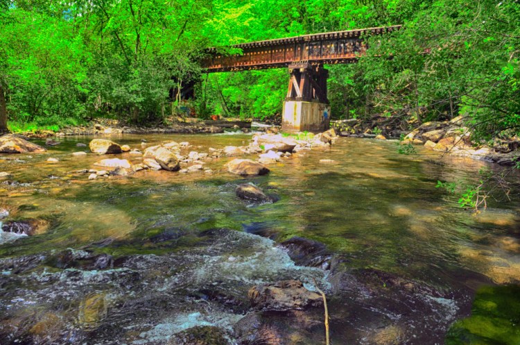 This June 30, 2016 photo shows Cobbosseecontee Stream flowing under a railroad trestle in Gardiner. There are plans to use the old trestle as part of a trail system along the stream.