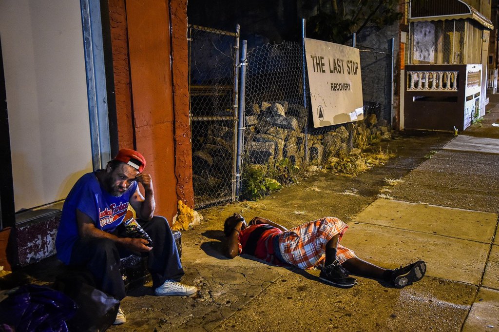 Art Gutierrez, 42, left, sits at a storefront in July, as another man sleeps on the sidewalk in the Kensington neighborhood of Philadelphia. Fentanyl has been creeping into the heroin supply there, leading to a rise in overdoses.