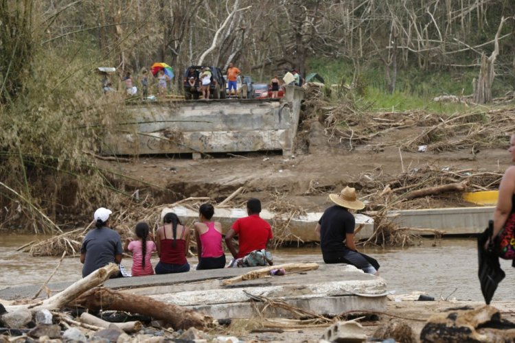 People sit on both sides of a destroyed bridge that crossed the San Lorenzo de Morovis River in Morovis, Puerto Rico. Maria has thrown Puerto Rico's already messy economic recovery plans into disarray.
