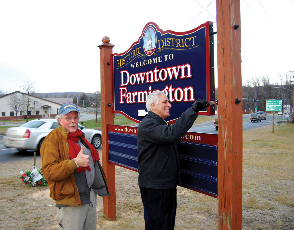 Mike Monahan gives a thumbs-up as Peter Judkins, president of Franklin Savings Bank, works on the Farmington Downtown Association sign. Monahan created many signs for the community and area businesses. He died March 26.