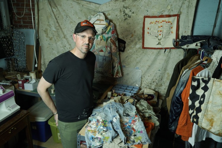 Jared DeSimio in the basement workspace at his home. DeSimio creates one-of-a-kind clothing from old garments, and also mends clothes, like the shorts of an artist friend of his seen on the table in the foreground.