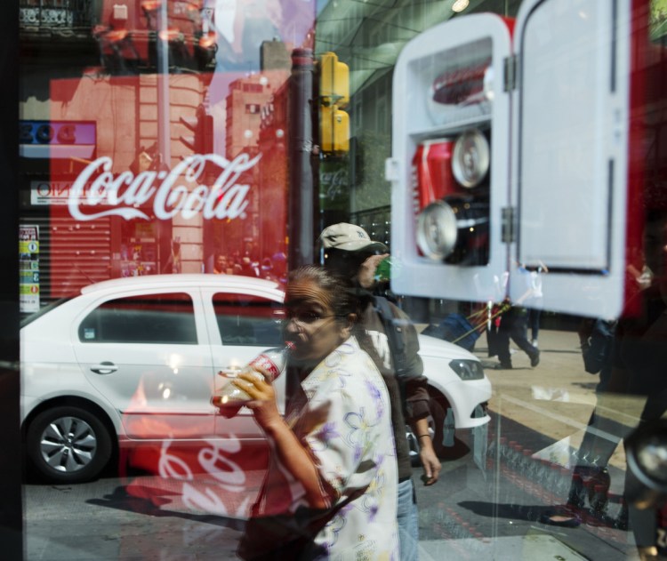 A woman is reflected in a Coca-Cola store window display as she drinks a Coke in Mexico City. In the southern city of Ciudad Altamirano, a store owner says a drug gang has seized control of soda distribution and raised prices.