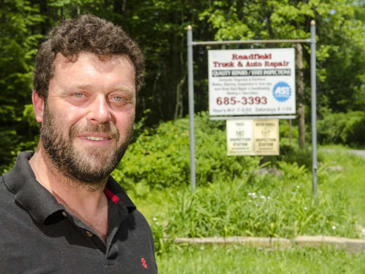 Matthew Curtis stands by his auto repair business sign Wednesday at the corner of North Road and Terrace Road in Readfield. Town officials call his property an illegal junkyard, which Curtis disputes.