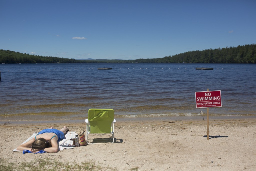 Christy Dow of Naples tans Saturday on Woods Pond Beach in Bridgton, next to one of the no-swimming signs that lined the sand. Swimming at the popular beach was banned when several people fell ill after being in the water, but officials reopened the swimming area Tuesday because tests showed the water is safe. 