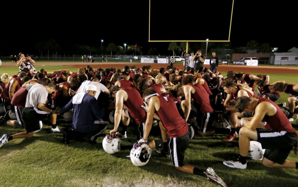 Members of the Marjory Stoneman Douglas High School football team pray together as they began practice for a new season just after midnight on Monday. Below, a coach wears a logo honoring the 17 lives lost in the February shooting.