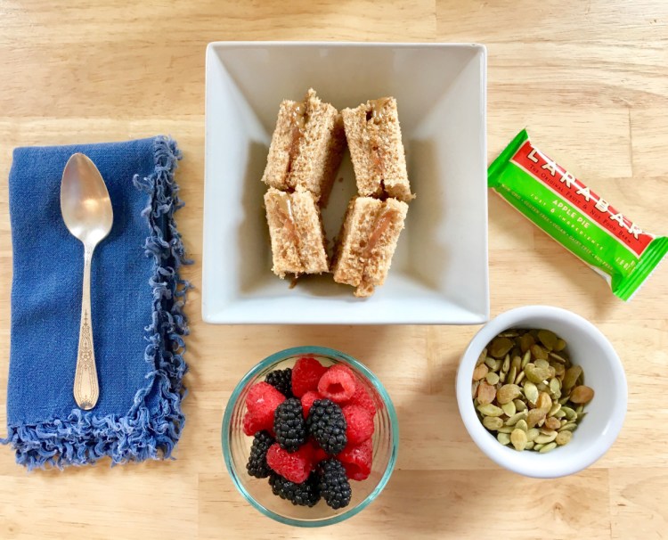 A simple vegan lunchbox meal includes sunbutter and jam sandwich squares, fresh mixed berries, roasted pumpkin seeds with golden raisins, and an apple pie Larabar.