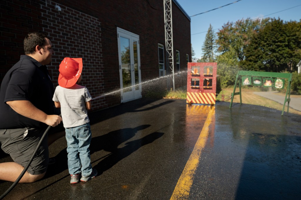 Winslow on-call firefighter Joel Cloutier, left, assists Jamison Emery, 3 of Winslow, in an exercise that teaches children to aim a water hose to put a fire out. Cloutier, who has worked with the Winslow Fire Department for one week, was taking part in an open house Saturday morning at the Winslow Fire Department.