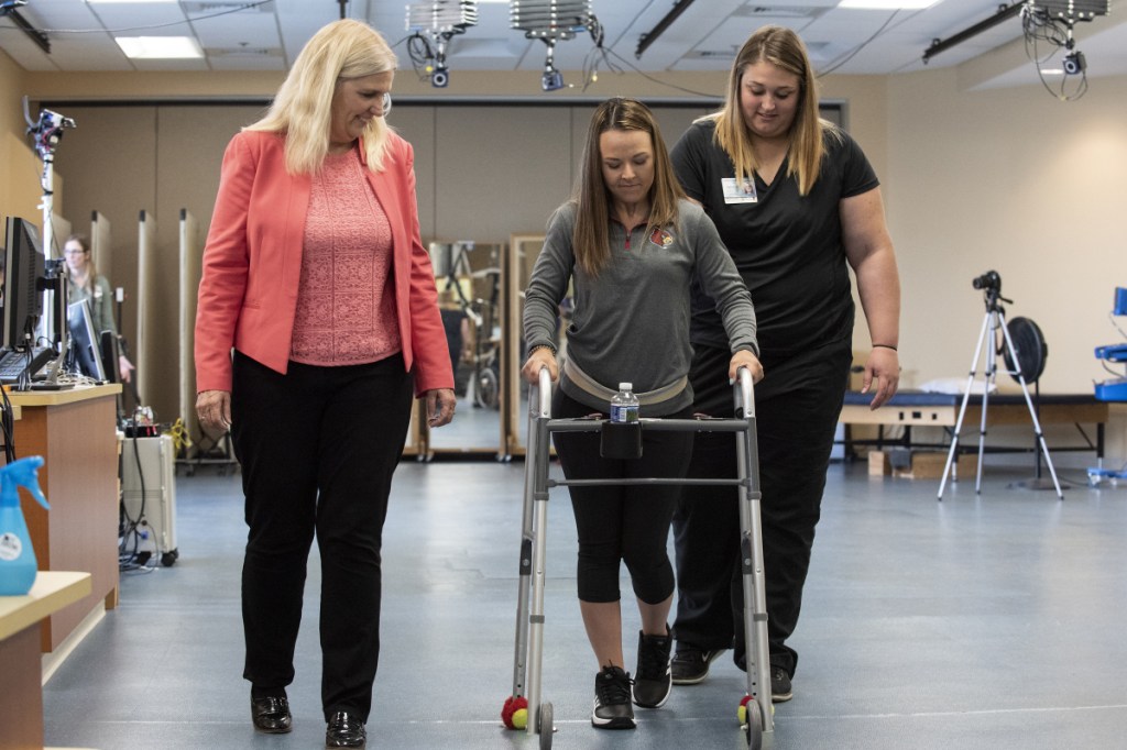 In this May 22, 2018 photo provided by the University of Louisville, Professor Susan Harkema watches as Kelly Thomas of Lecanto, Fla., practices walking with the help of a walker. "Recovery can happen if you have the right circumstances," says Harkema, who co-authored a report on the use of electrical implants to stimulate the spinal cord. (Tom Fougerousse/University of Louisville via AP)