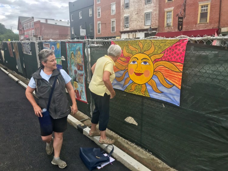 Deborah Fahy, left, Harlow Gallery executive director, and Nancy Bischoff, a member of the Down with the Crown Committee, hang a mural near the corner of Central and Water streets in Hallowell. Both women are involved in the Murals of the Fence project that hung artwork along construction fencing along Water Street.