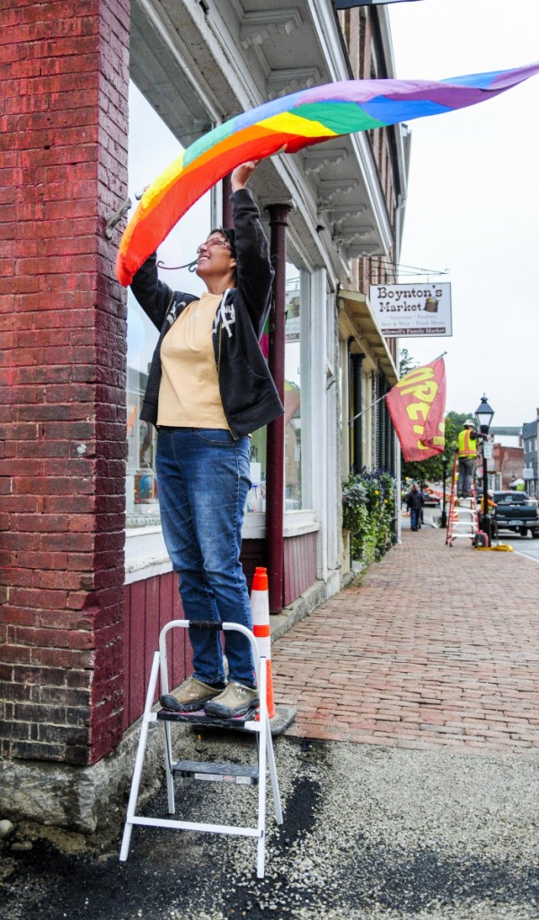 Standing on a stepladder, Malley Weber hangs a flag outside of her shop, Hallowell Clay Works, on Tuesday on Water Street in downtown Hallowell. She said that when workers finish installing the new brick sidewalks at the corner of Water and Union streets she should be able to reach the flag mount without a stepladder again.