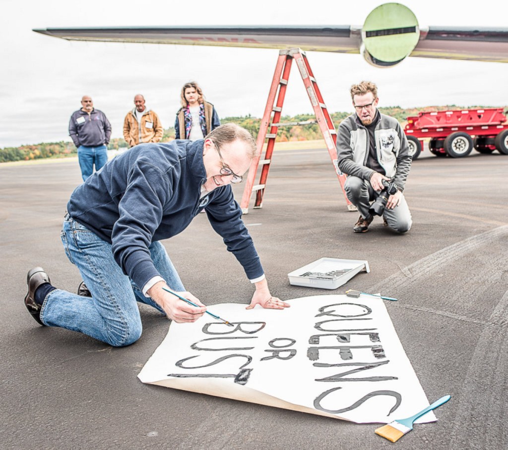 MCR CEO Tyler Morse paints a sign, reading "Queens or bust," before hanging it on the back of the 150-foot fuselage. Visitors to the plane-turned-cocktail lounge will "feel like you walked into 1962," he said.