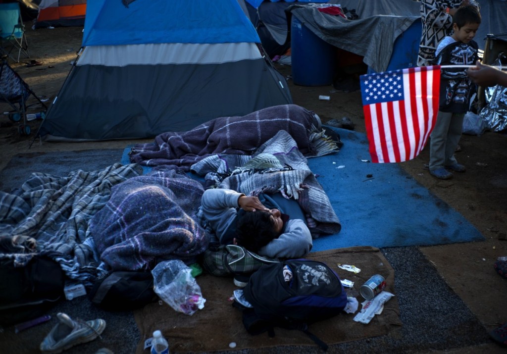 A migrant holding a U.S. flag, right, speaks with others waking up at the Benito Juarez Sports Center that's serving as a temporary shelter in Tijuana, Mexico, early Monday. The mayor of Tijuana has declared a humanitarian crisis in his border city and says that he has asked the United Nations for aid to deal with thousands of Central American migrants who have arrived in the city.