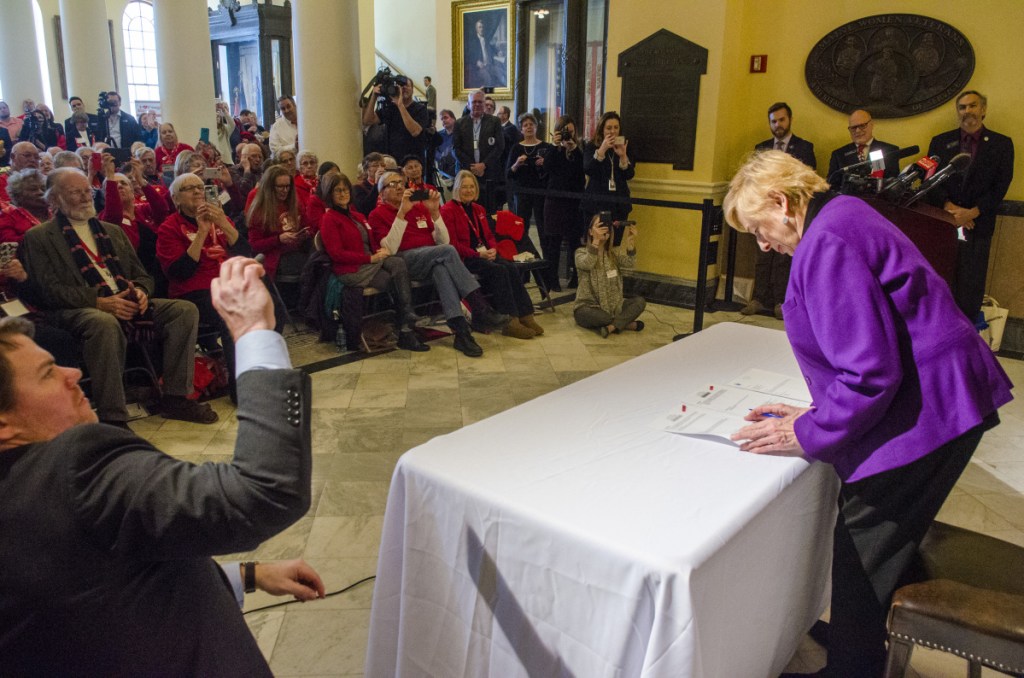Spectators, many wearing red AARP shirts, watch as Gov. Janet Mills signs bonds for affordable senior housing on Tuesday in the Maine State House Hall of Flags in Augusta. Mills' predecessor, Republican Gov. Paul LePage, had refused to release the bonds for more than three years.