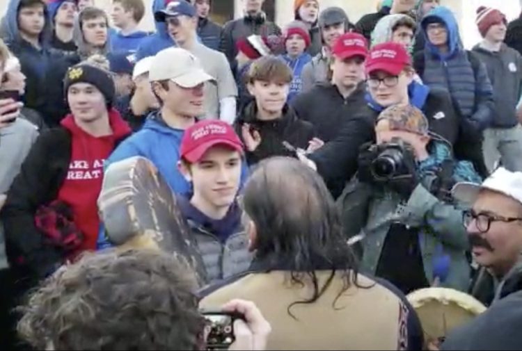 In this Friday, Jan. 18, 2019 image made from video provided by the Survival Media Agency, a teenager wearing a "Make America Great Again" hat, center left, stands in front of an elderly Native American singing and playing a drum in Washington. The Roman Catholic Diocese of Covington in Kentucky is looking into this and other videos that show youths, possibly from the diocese's all-male Covington Catholic High School, mocking Native Americans at a rally in Washington. 