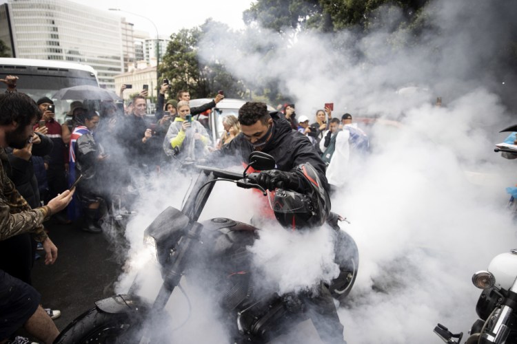 Protesters watch as a man spins the tire on his motorcycle in wet conditions as they demonstrate their opposition to coronavirus vaccine mandates at Parliament in Wellington, New Zealand, Saturday. The protest began when a convoy of trucks and cars drove to Parliament from around the nation, inspired by protests in Canada. (George Heard/NZME via AP)